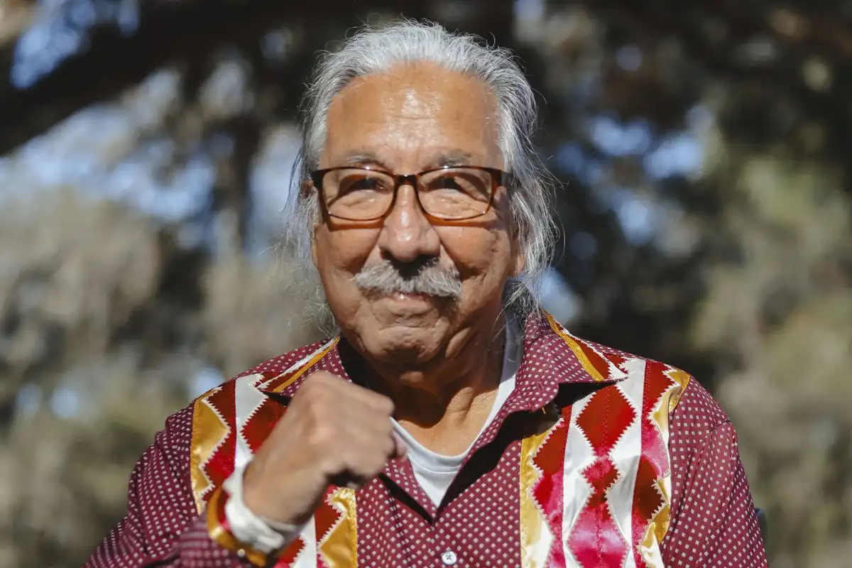 An elderly man with white hair, glasses, and a mustache wears a red, white, and gold patterned shirt while making a fist…