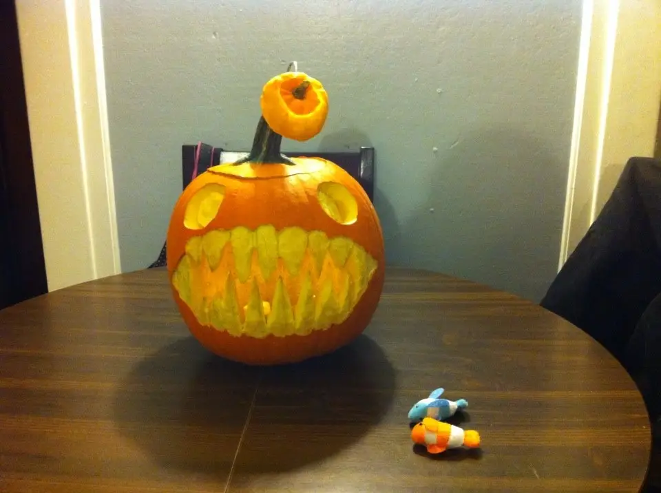 a carved jack-o'-lantern with a wide, jagged mouth and hollow eyes sits on a wooden table, its stem topped with a small…