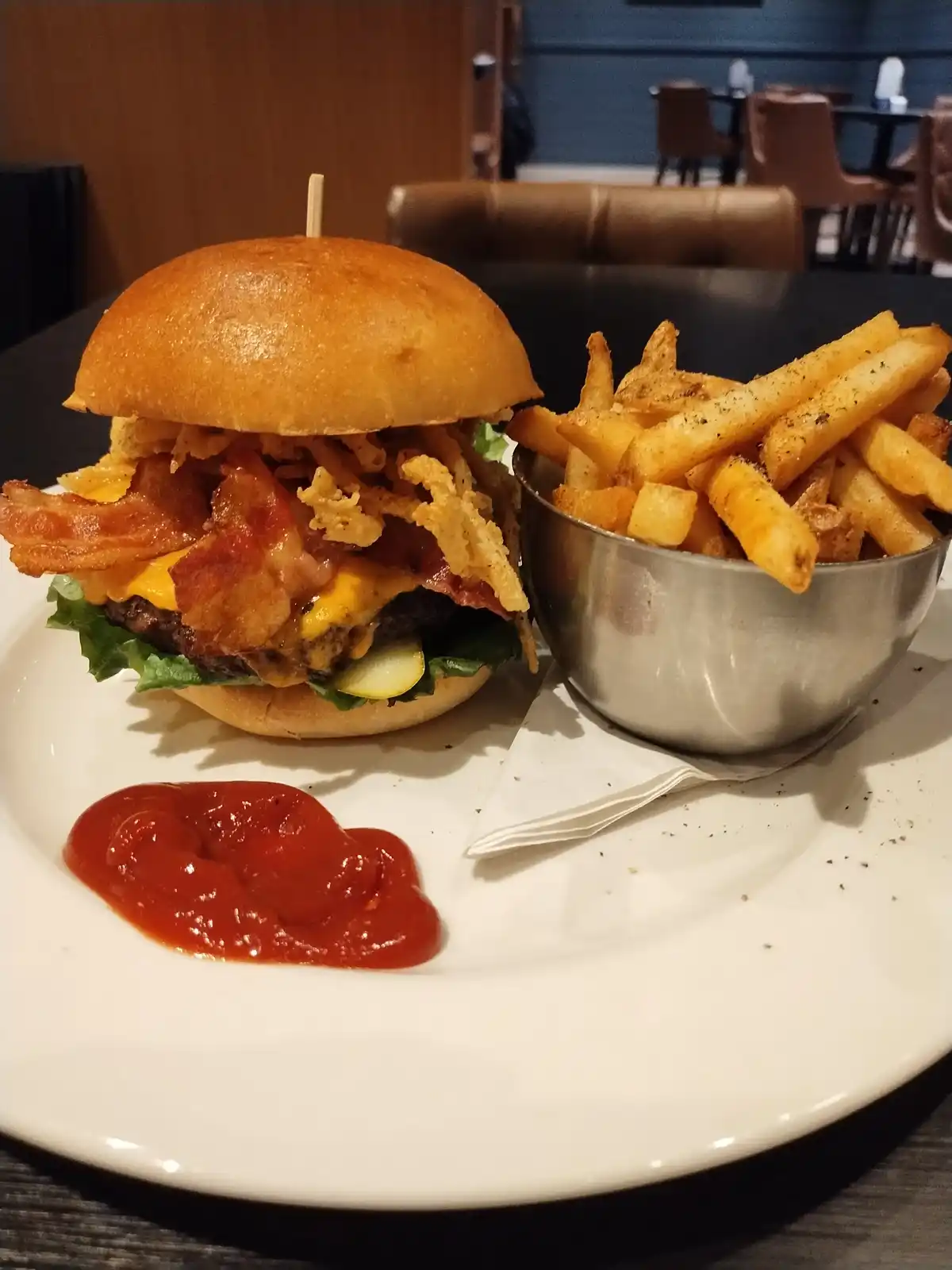 A close-up photo of a heavily loaded burger with bacon, melted cheese, lettuce, and crispy onion rings, held together by a…