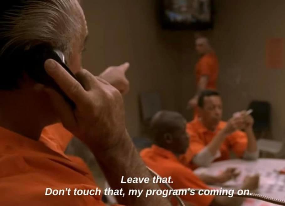 A man in an orange prison jumpsuit is on the phone, pointing at something off-screen while other inmates sit nearby. The…