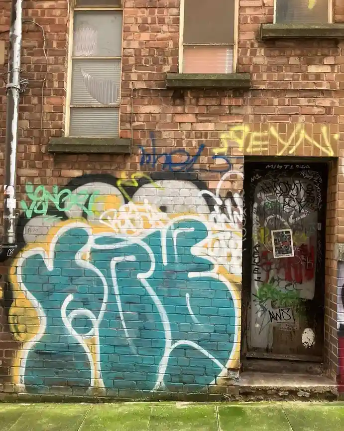 A brick wall covered in colorful graffiti, including a large blue and yellow abstract mural, with a heavily tagged door to…
