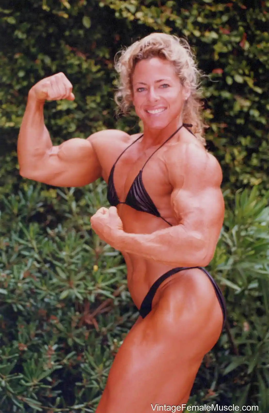 A muscular woman with blonde hair, wearing a black bikini, poses outdoors in front of green bushes, flexing both arms to…