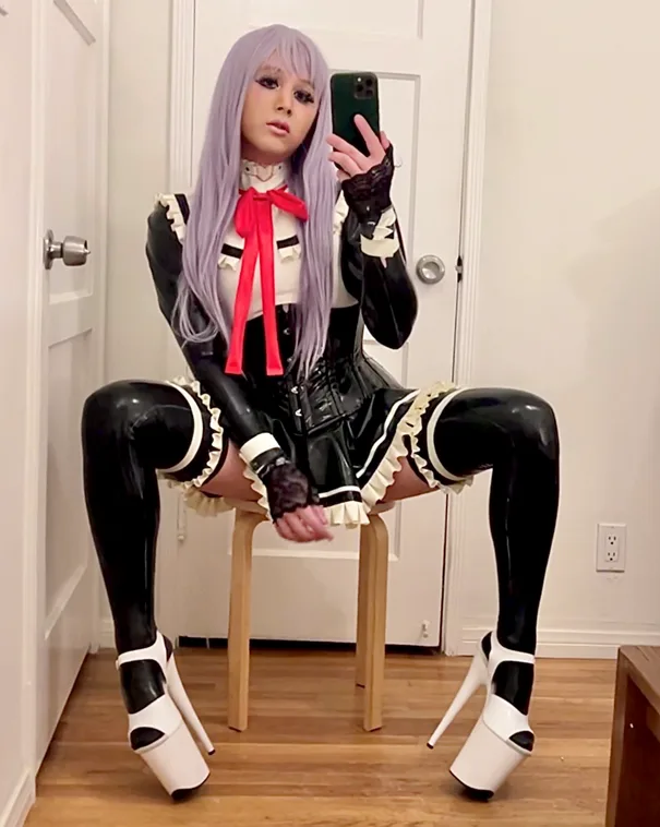 A person with long lavender hair takes a mirror selfie while wearing a black and white maid outfit with a red bow, black…