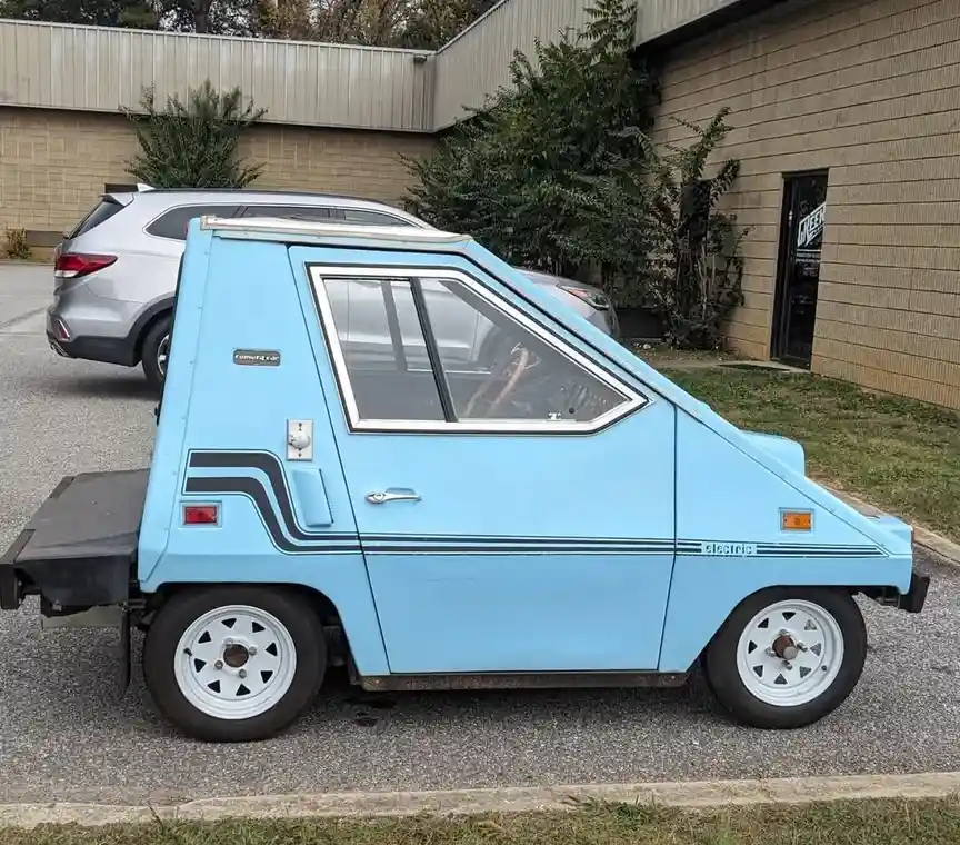 A light blue, triangular-shaped, electric 'Comuta Car' is parked on asphalt next to a modern silver SUV. The car has a very…