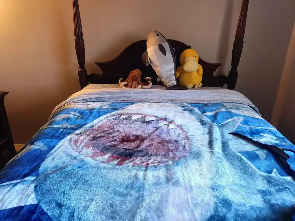 A bed with a shark-themed blanket featuring a large open mouth, has three plush toys on the headboard: a brown octopus, a…