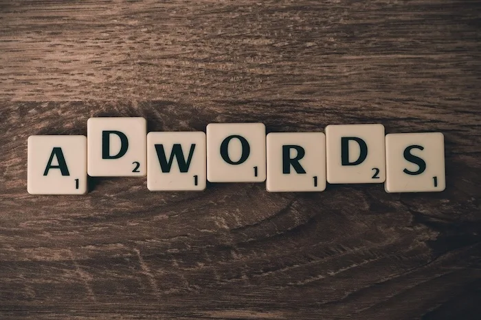 The word 'ADWORDS' is spelled out using white Scrabble tiles with black lettering on a dark wooden surface. Each tile has a…