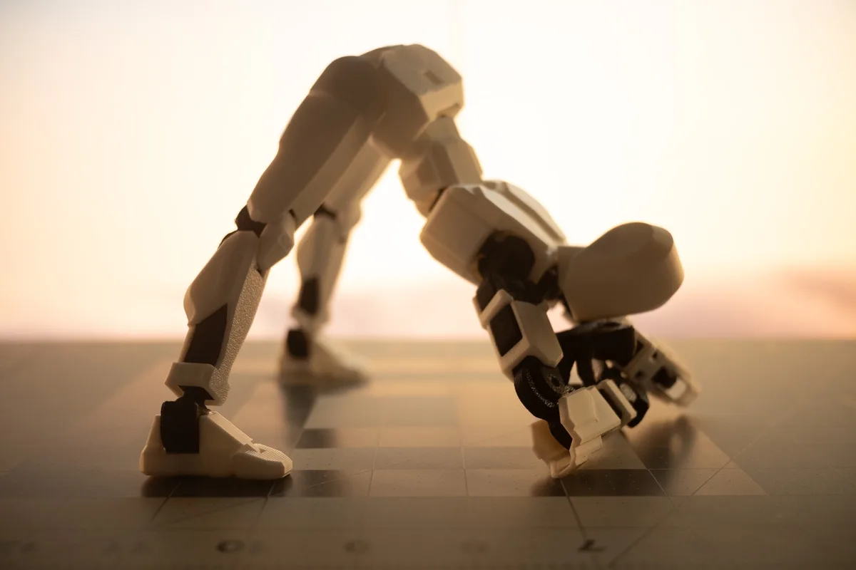 A white and black articulated robot figurine is bent over in a yoga-like pose on a checkered surface, with a warm, hazy…