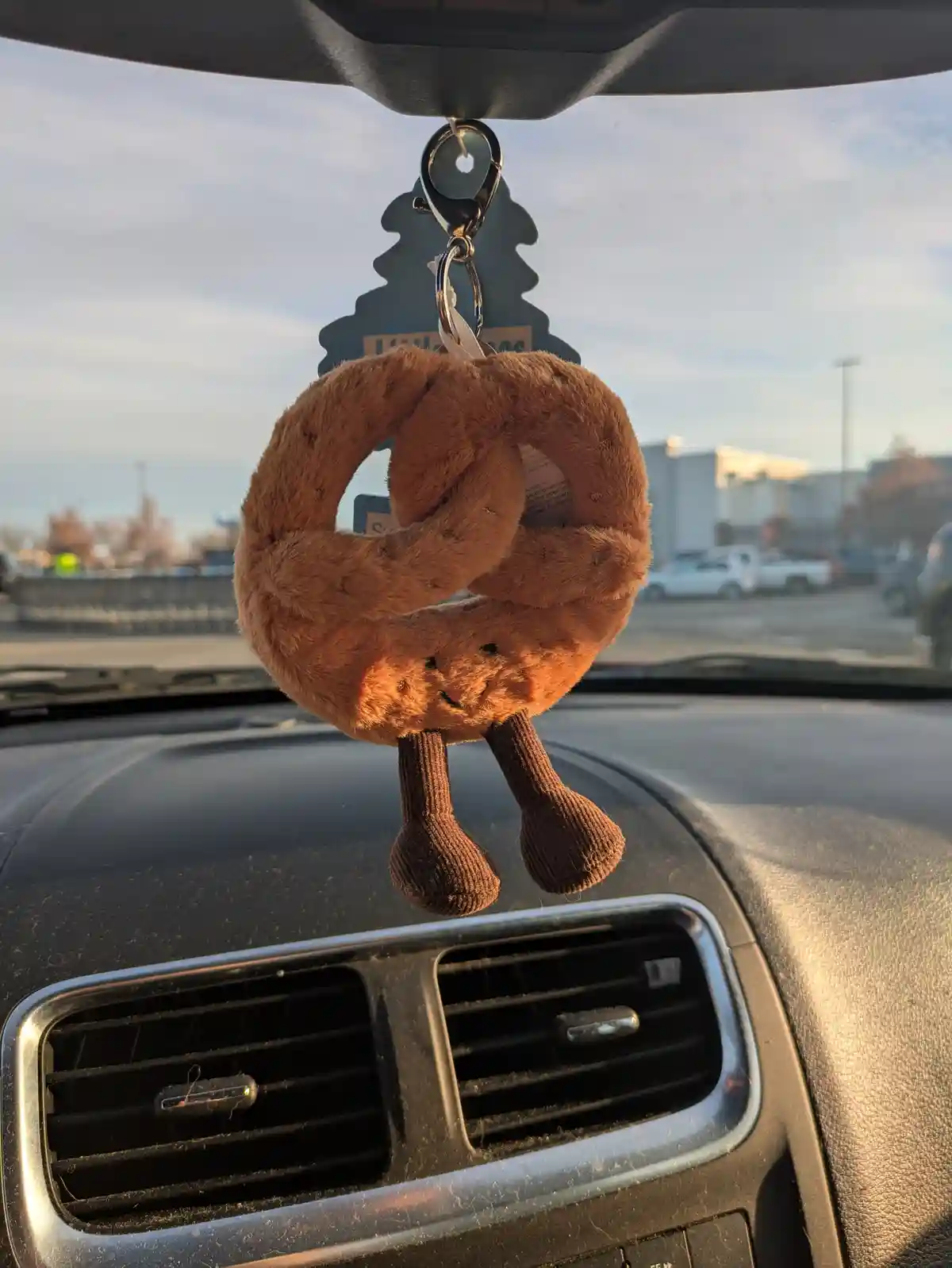 A plush toy shaped like a pretzel with a smiling face and knitted legs hangs from a car's rearview mirror, attached to a car…