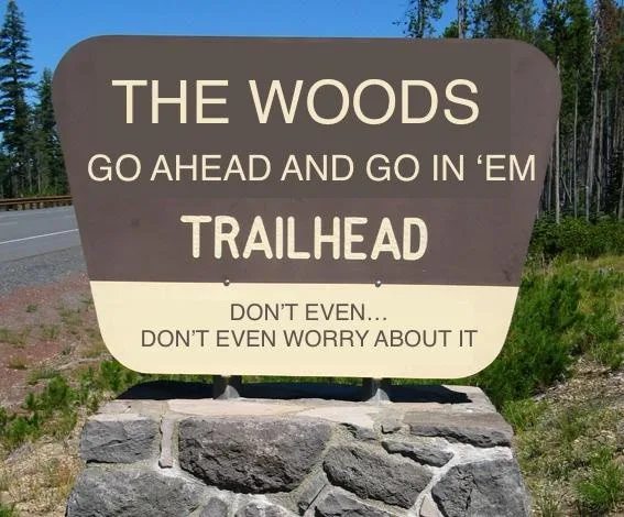 A brown and beige trailhead sign reads 'THE WOODS GO AHEAD AND GO IN 'EM TRAILHEAD' with a second section below saying…