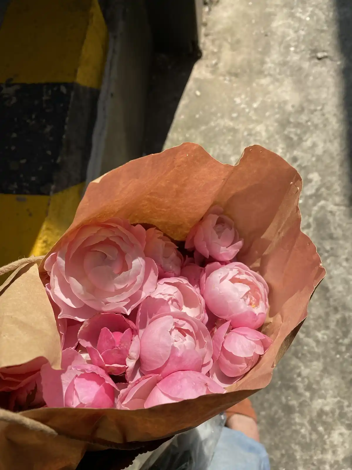 A close-up photo shows a bouquet of pink roses and peonies wrapped in brown paper, held outdoors near a yellow-and-black…