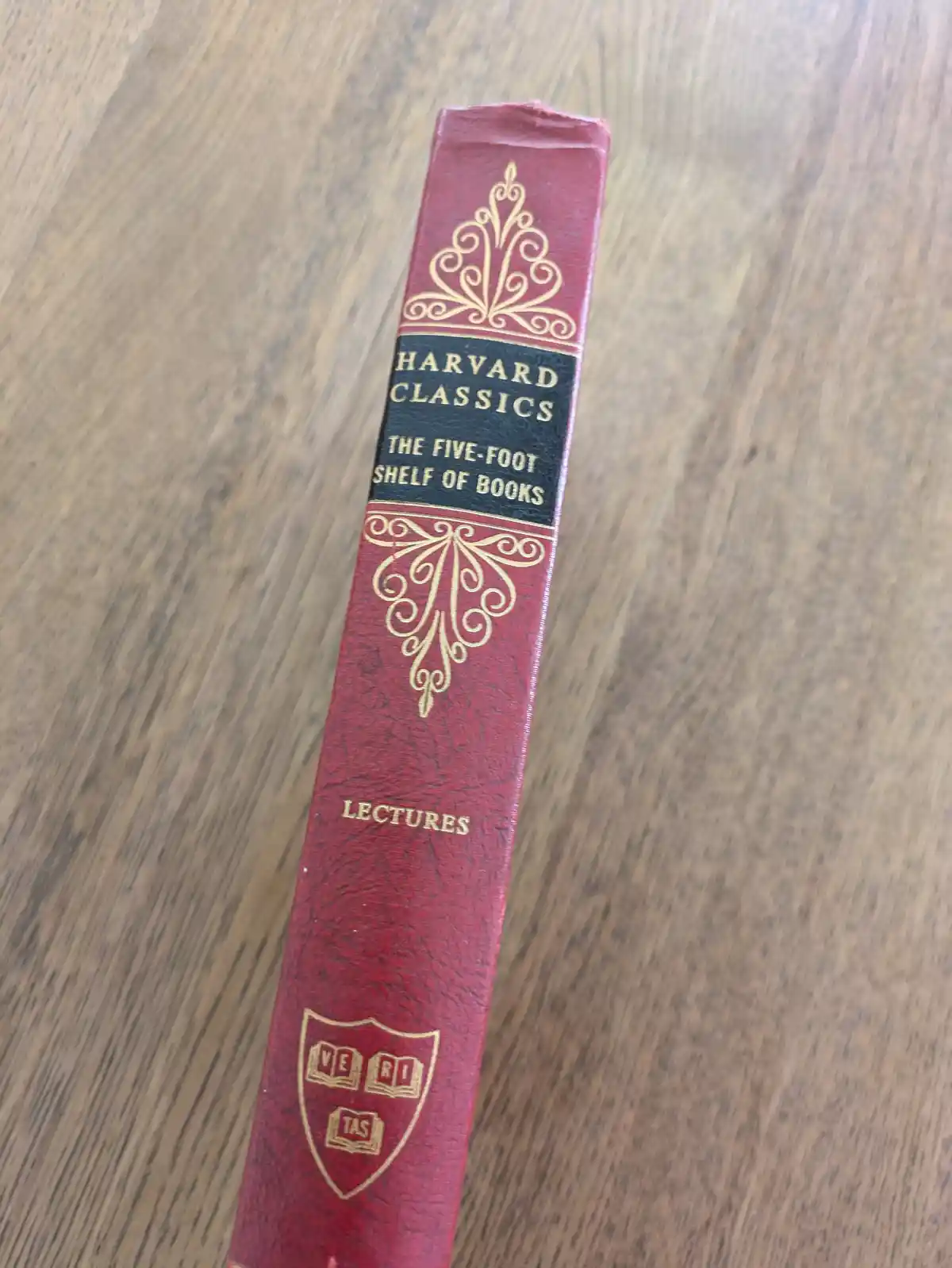 A close-up of a red leather-bound book spine on a wooden surface, showing gold embossed text and decorative flourishes. The…