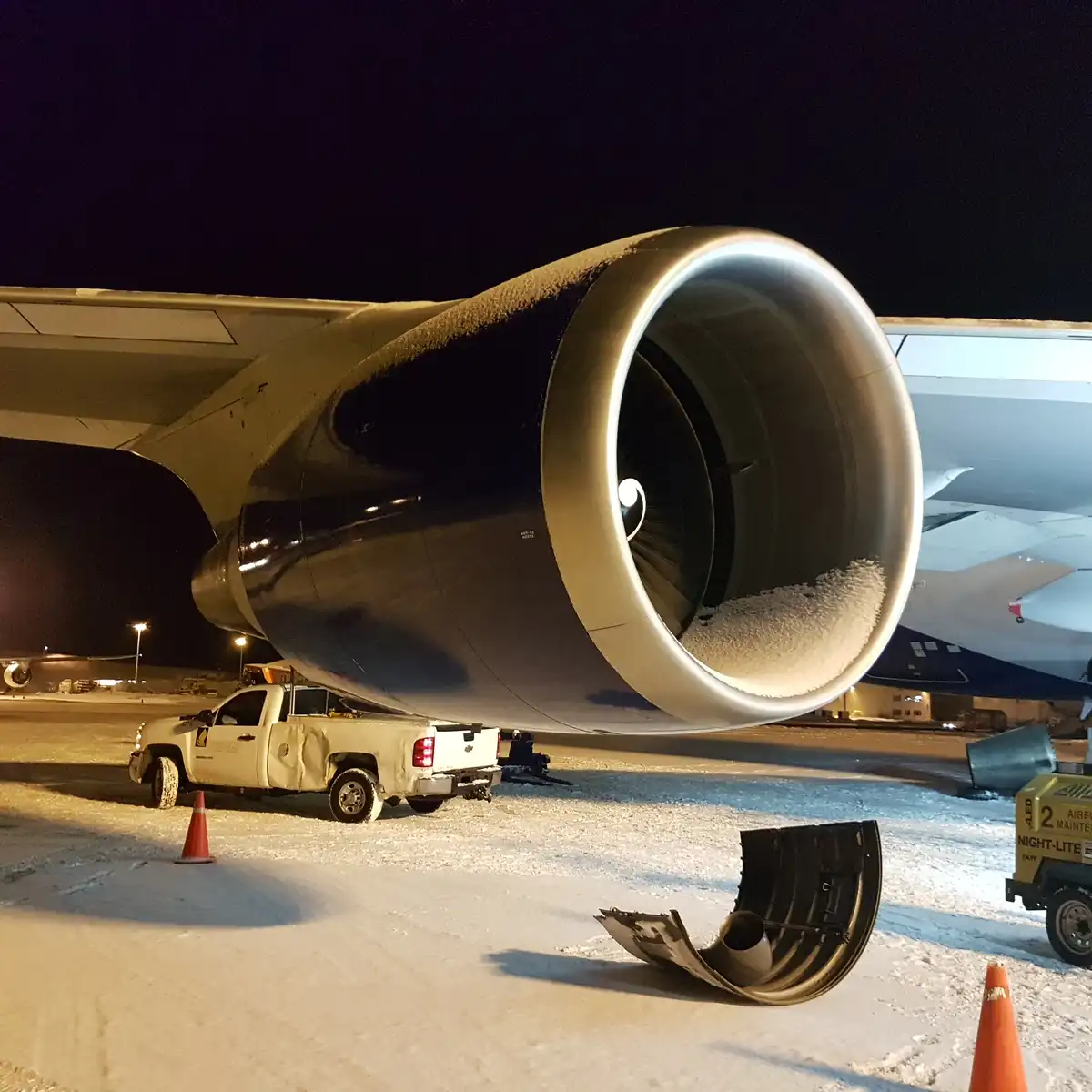 A close-up of a large airplane engine at night, covered in snow, with a damaged engine cowling lying on the snowy tarmac…