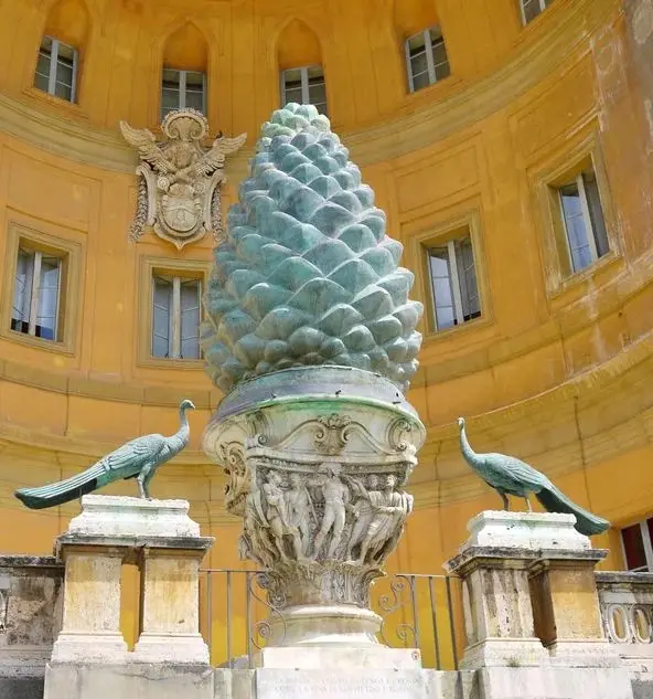 a large, ornate bronze pinecone sculpture sits atop a decorative pedestal in front of a yellow building with arched windows…
