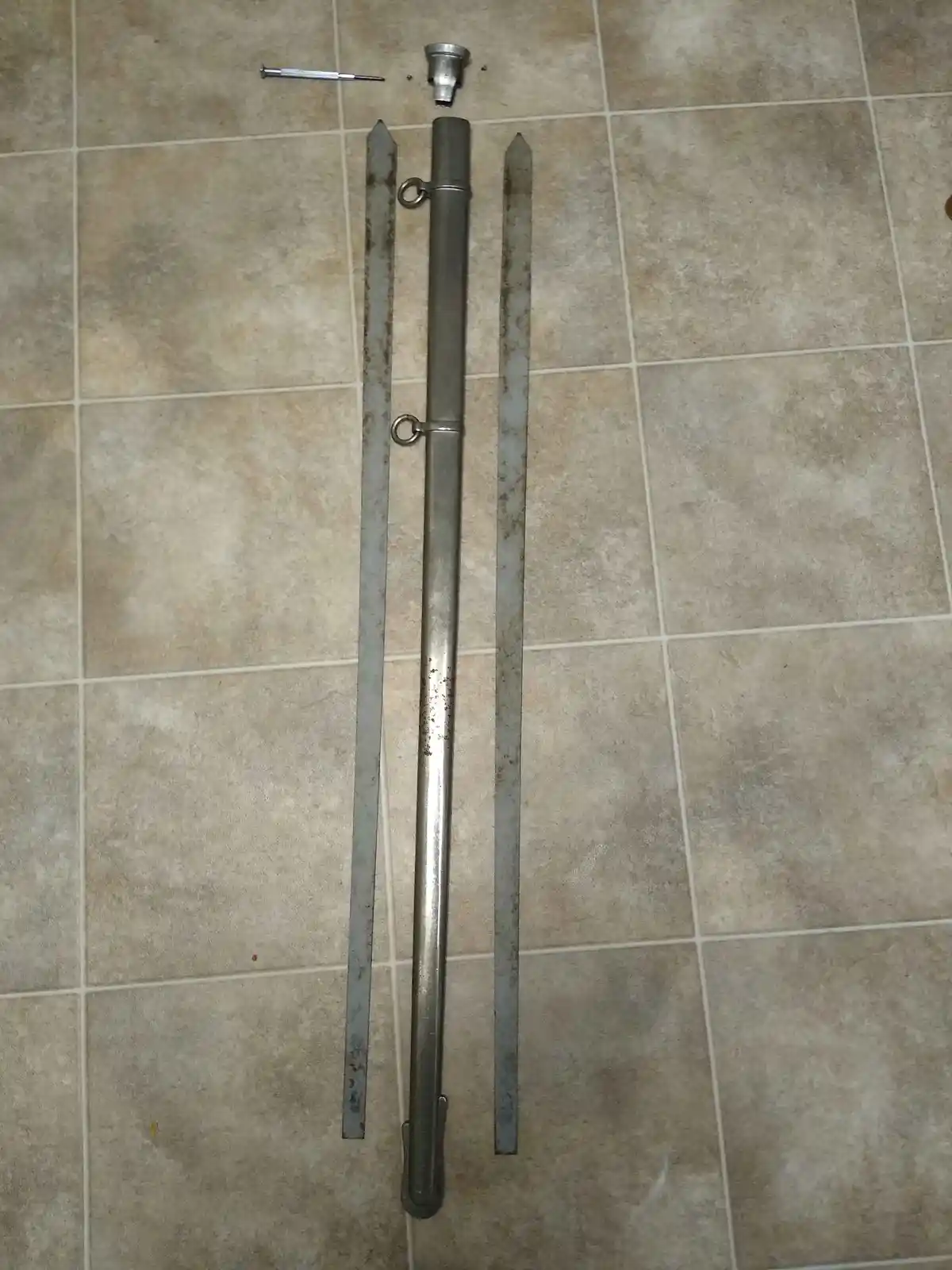 A disassembled metal object, resembling a vintage or industrial tool, lies on a tiled floor. It consists of a central long…