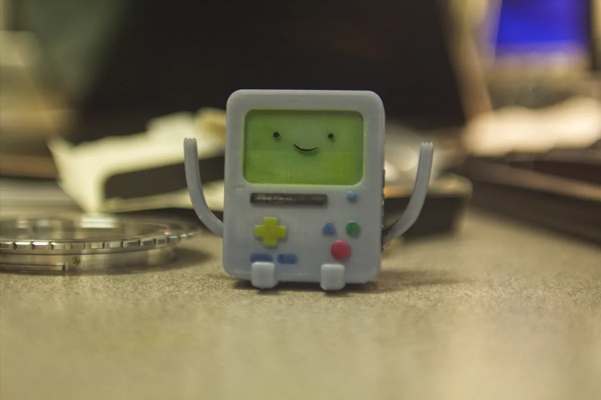 A small, 3D-printed figurine of the character BMO from Adventure Time, resembling a Game Boy with a smiling green screen…