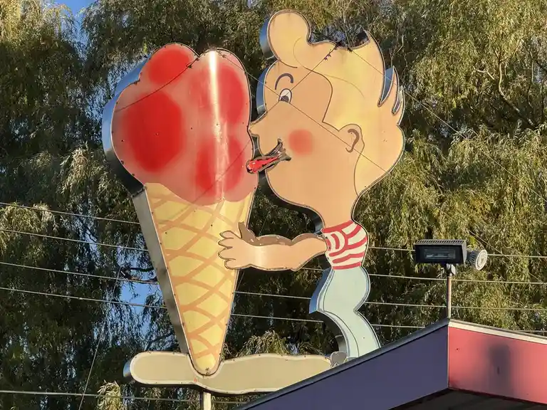 A large, weathered, vintage-style metal sign depicts a cartoon boy in a red-and-white striped shirt licking a giant red ice…