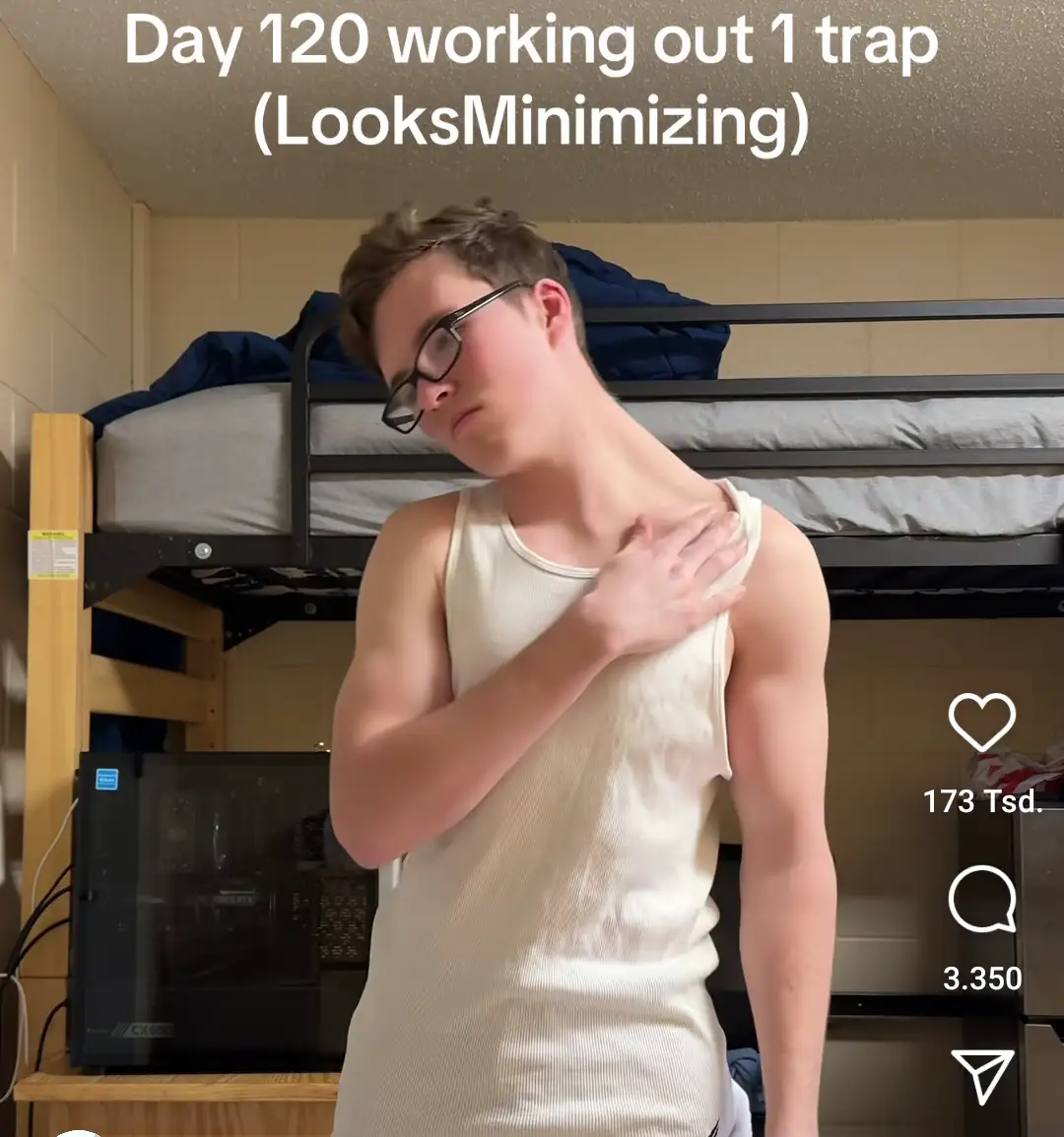 A young man in glasses and a white tank top stands in a dorm room with bunk beds behind him, touching his shoulder with a…