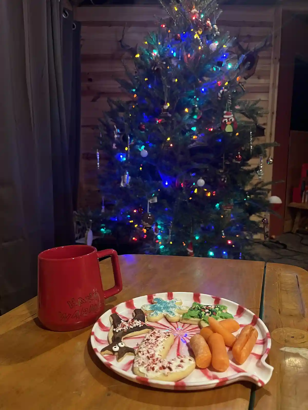 A red mug labeled 'HAND WARMER' sits next to a plate of Christmas cookies and baby carrots on a wooden table, with a…