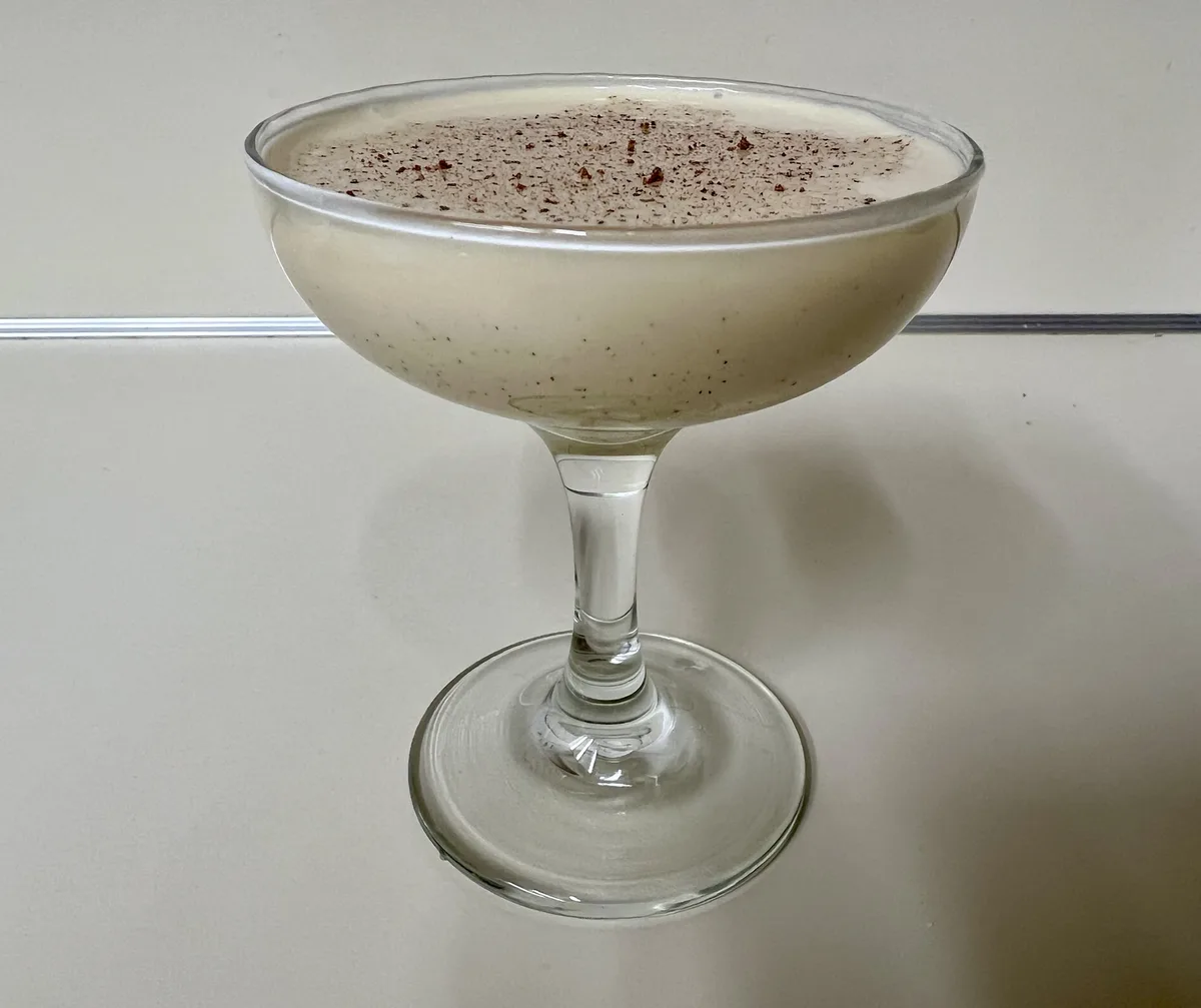 A creamy, light-brown beverage in a stemmed coupe glass, topped with a sprinkle of reddish-brown spice, sits on a plain…