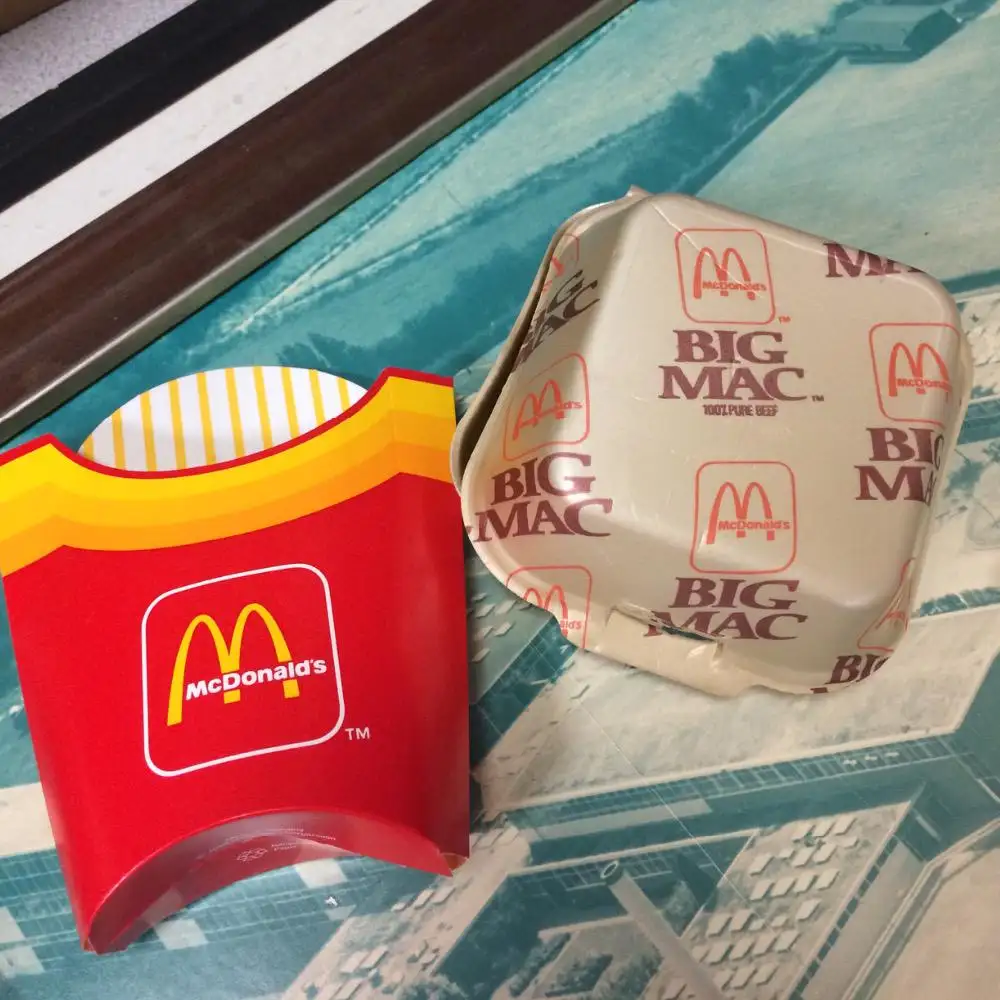 A red McDonald's french fry container sits next to a crumpled, beige Big Mac burger box, both featuring the McDonald's logo…