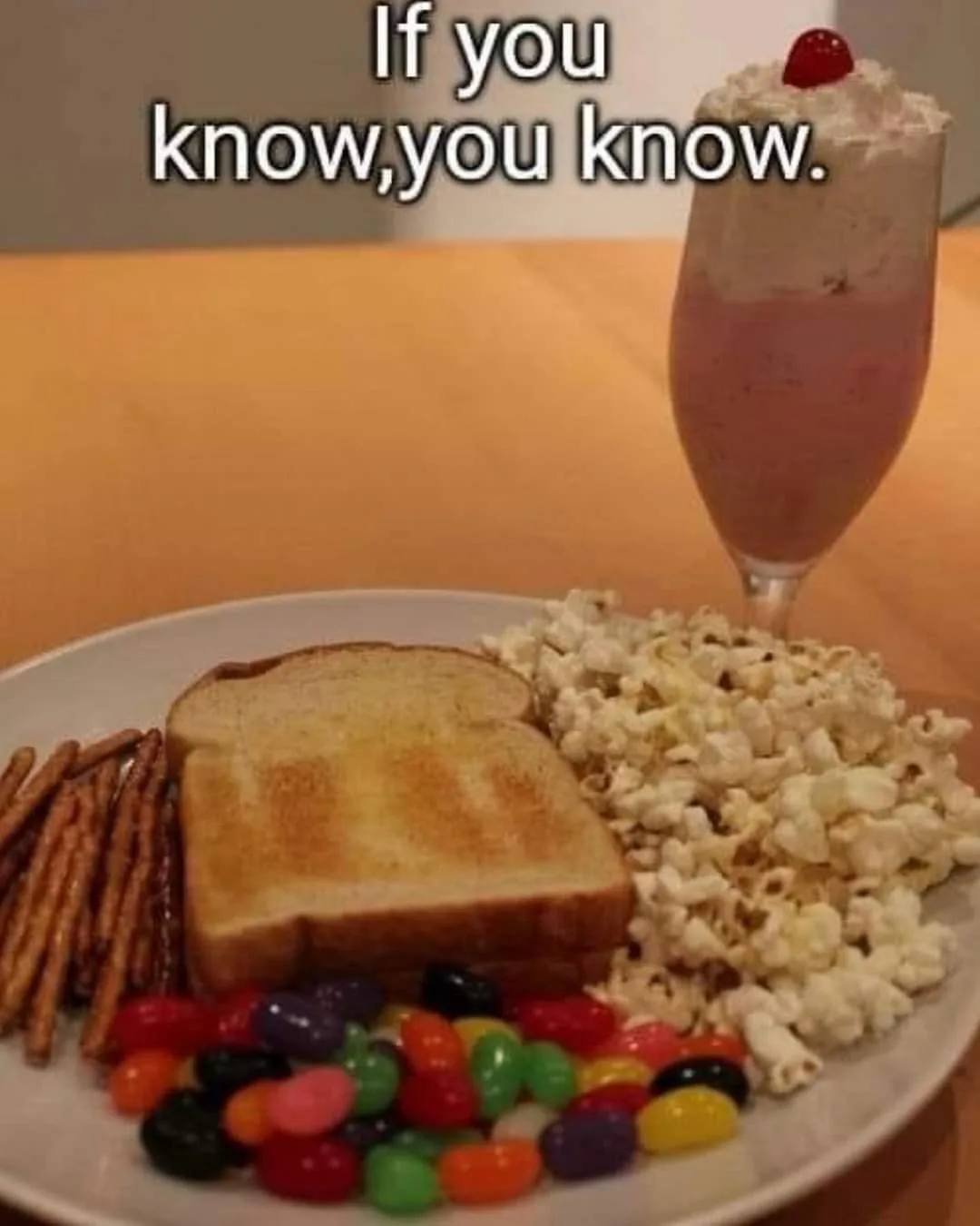 A plate of snacks including toast, pretzels, popcorn, and colorful jelly beans sits next to a pink milkshake topped with…