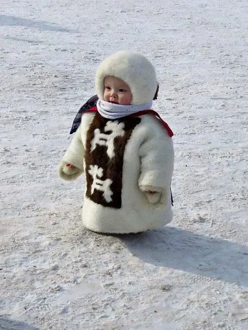 A baby dressed in a thick, white, furry winter coat with a hood and a scarf stands in a snowy field. The coat has a brown…