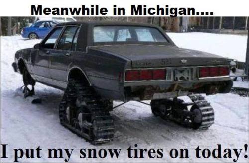 A dark gray sedan is parked in a snowy yard with tank treads instead of wheels, humorously implying it's 'snow tires'. The…