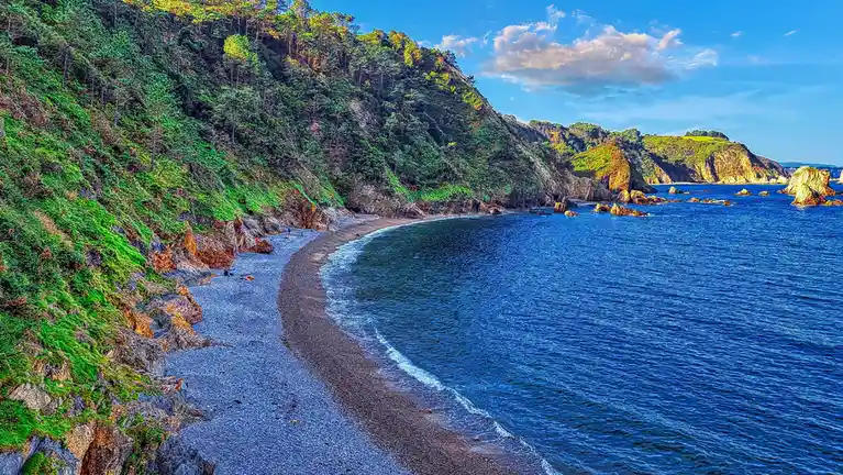 A vibrant, high-contrast photo of a secluded cove with a pebble beach, turquoise water, and forested cliffs under a partly…