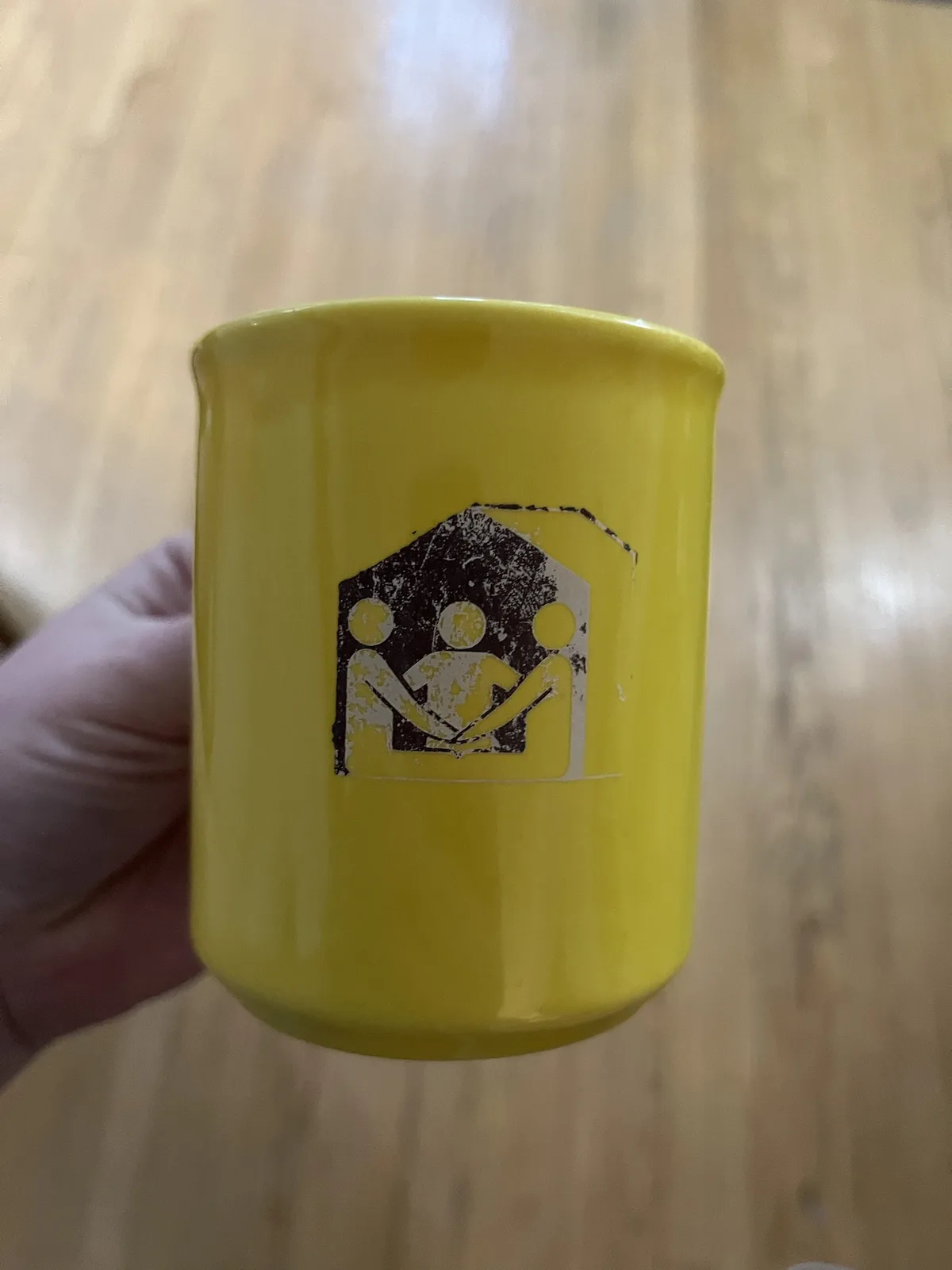 A hand holds a bright yellow mug against a wooden floor background. The mug features a worn, black-and-yellow graphic of…