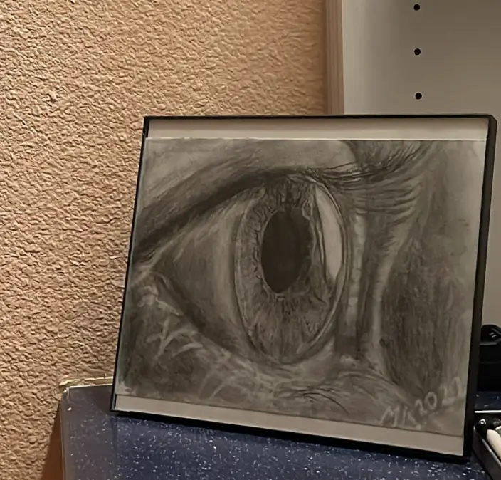 A framed, highly detailed pencil drawing of a human eye sits on a speckled blue surface against a textured beige wall. The…