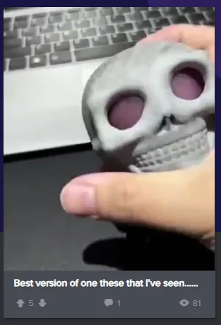 A hand holds a small, gray 3D-printed skull figurine with large, dark purple eye sockets and a visible toothy grin. In the…