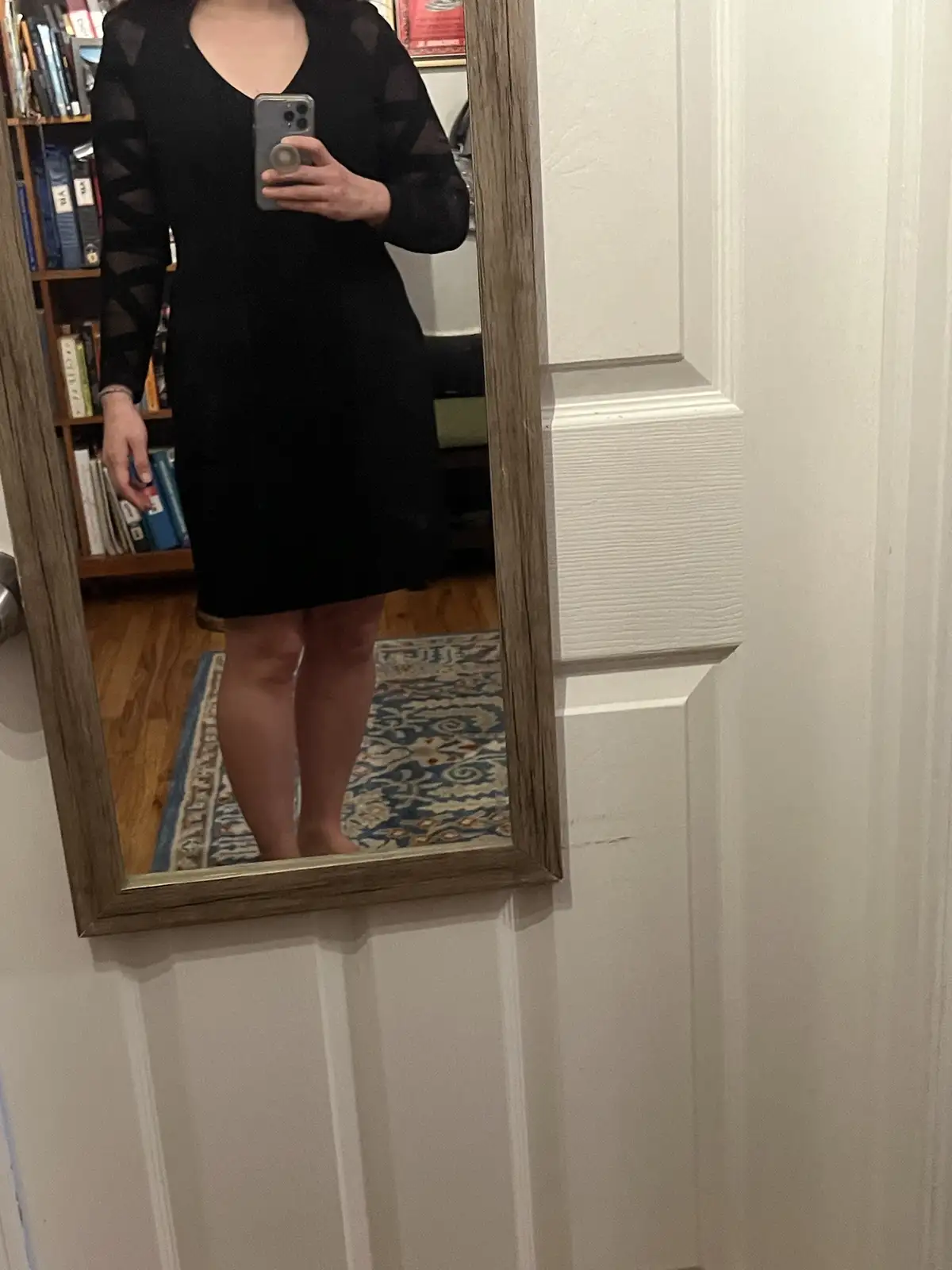 A person takes a mirror selfie wearing a black dress with sheer geometric-patterned sleeves, standing barefoot on a…