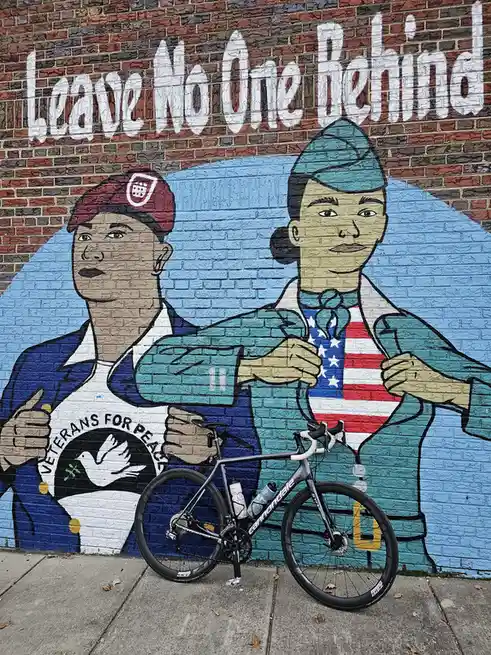 A bicycle is parked in front of a mural depicting two veterans, one wearing a 'Veterans for Peace' t-shirt and the other…