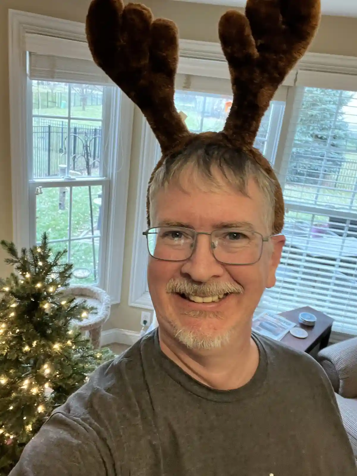 A smiling man with glasses and a mustache wears brown plush moose antlers indoors near a lit Christmas tree. The overlaid…