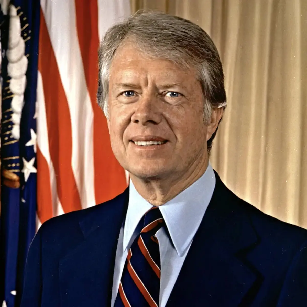 A formal portrait of former U.S. President Jimmy Carter, smiling, wearing a dark suit and striped tie, standing in front of…