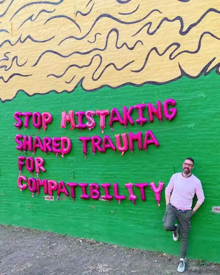 A man in a pink shirt and glasses leans casually against a brightly painted wall. The wall has a green lower half and a…