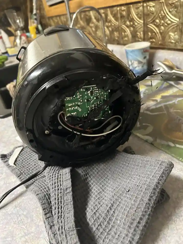 A black kitchen blender is shown with its bottom casing ripped open, revealing a green circuit board and wires covered in…