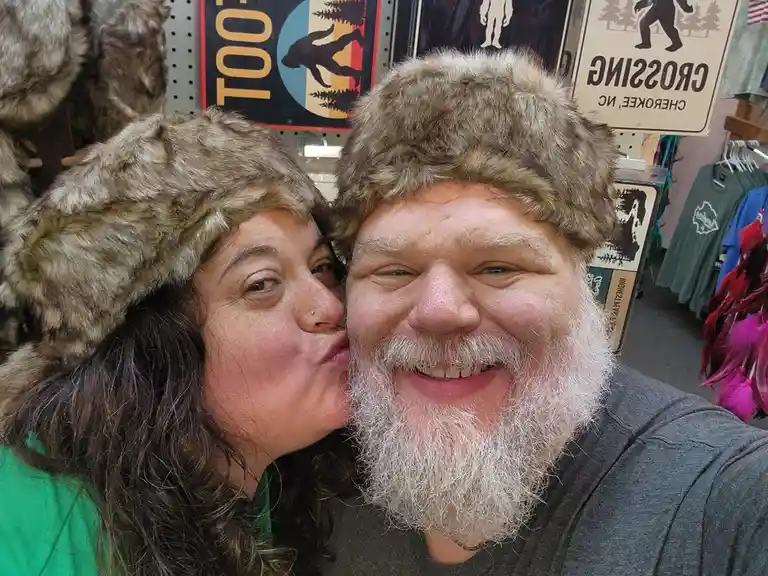 A selfie shows a smiling man with a white beard and a woman kissing his cheek, both wearing furry hats. Behind them are…