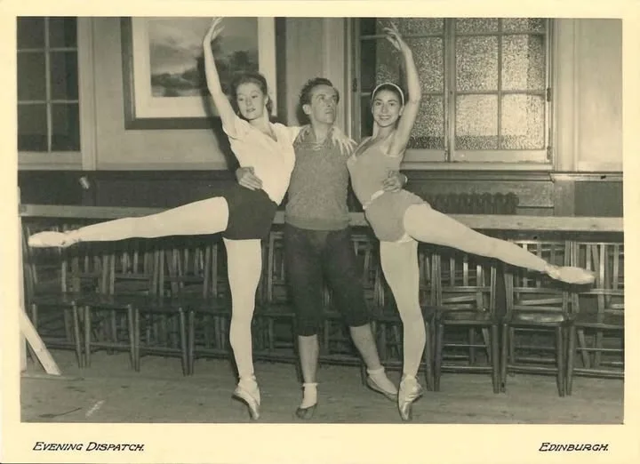 A vintage black-and-white photo shows a man standing between two female ballet dancers in mid-pose, each with one leg…