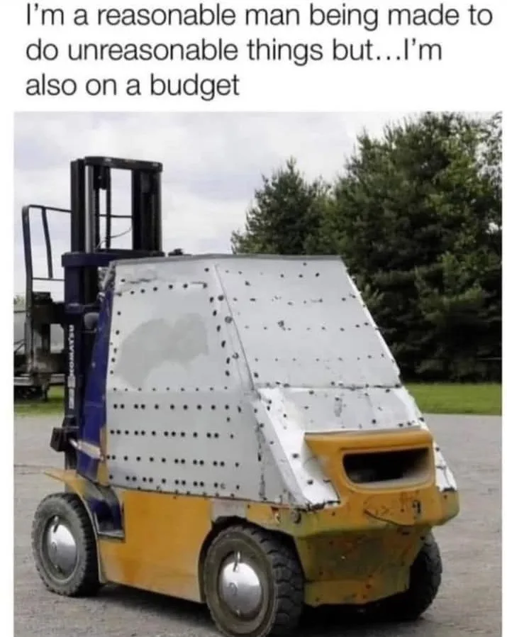 A yellow and blue forklift with a makeshift, riveted metal shield covering its front cab, parked outdoors with trees in the…