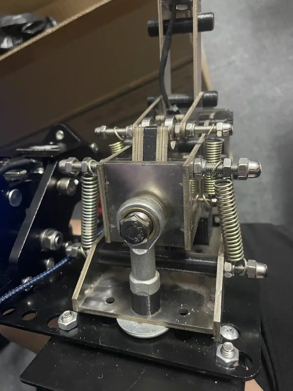 A close-up photo of a complex, homemade mechanical device made of metal, springs, bolts, and wires, mounted on a black…