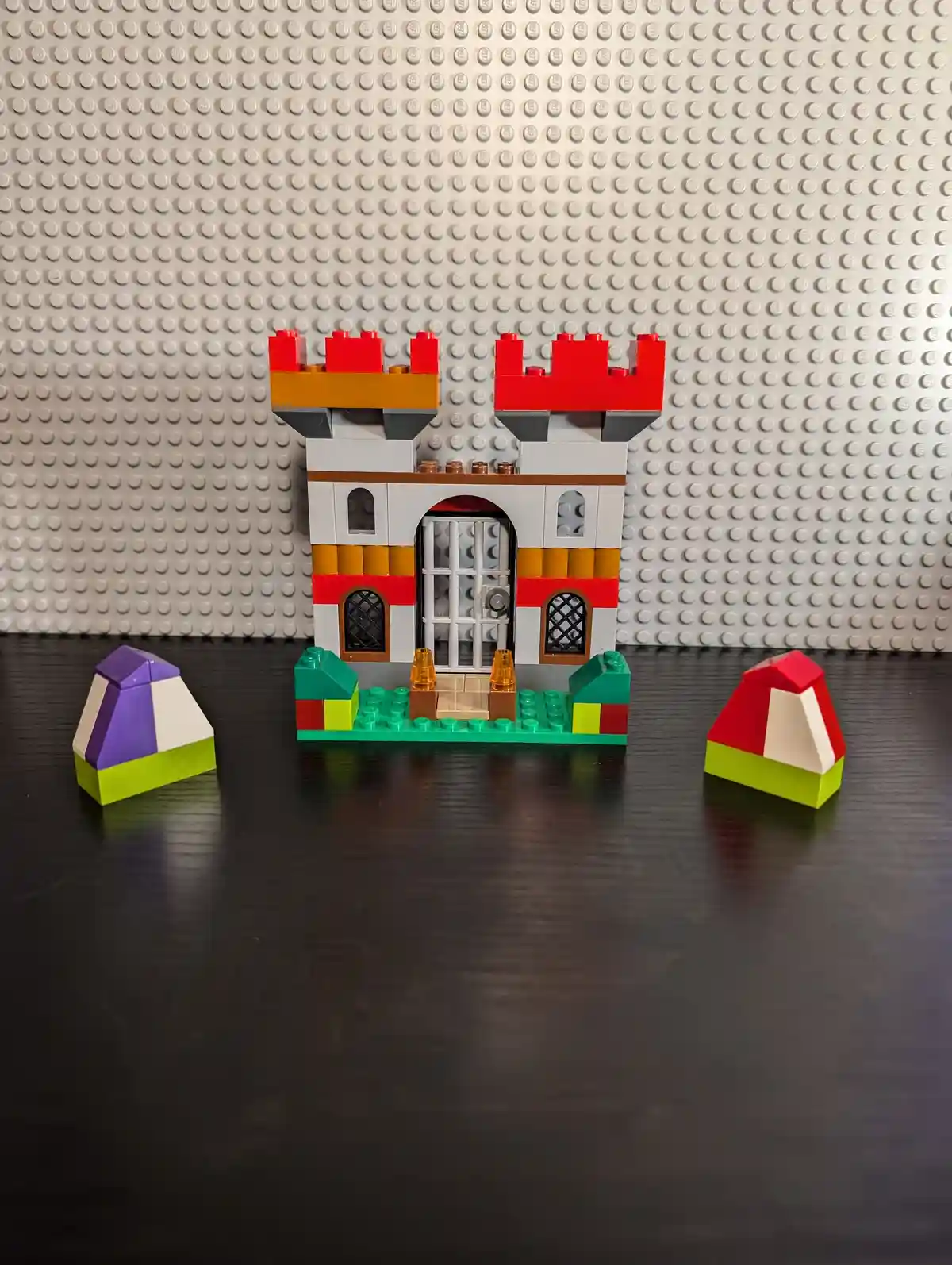 A small LEGO castle with red turrets and barred doors sits on a green baseplate, flanked by two colorful, incomplete LEGO…
