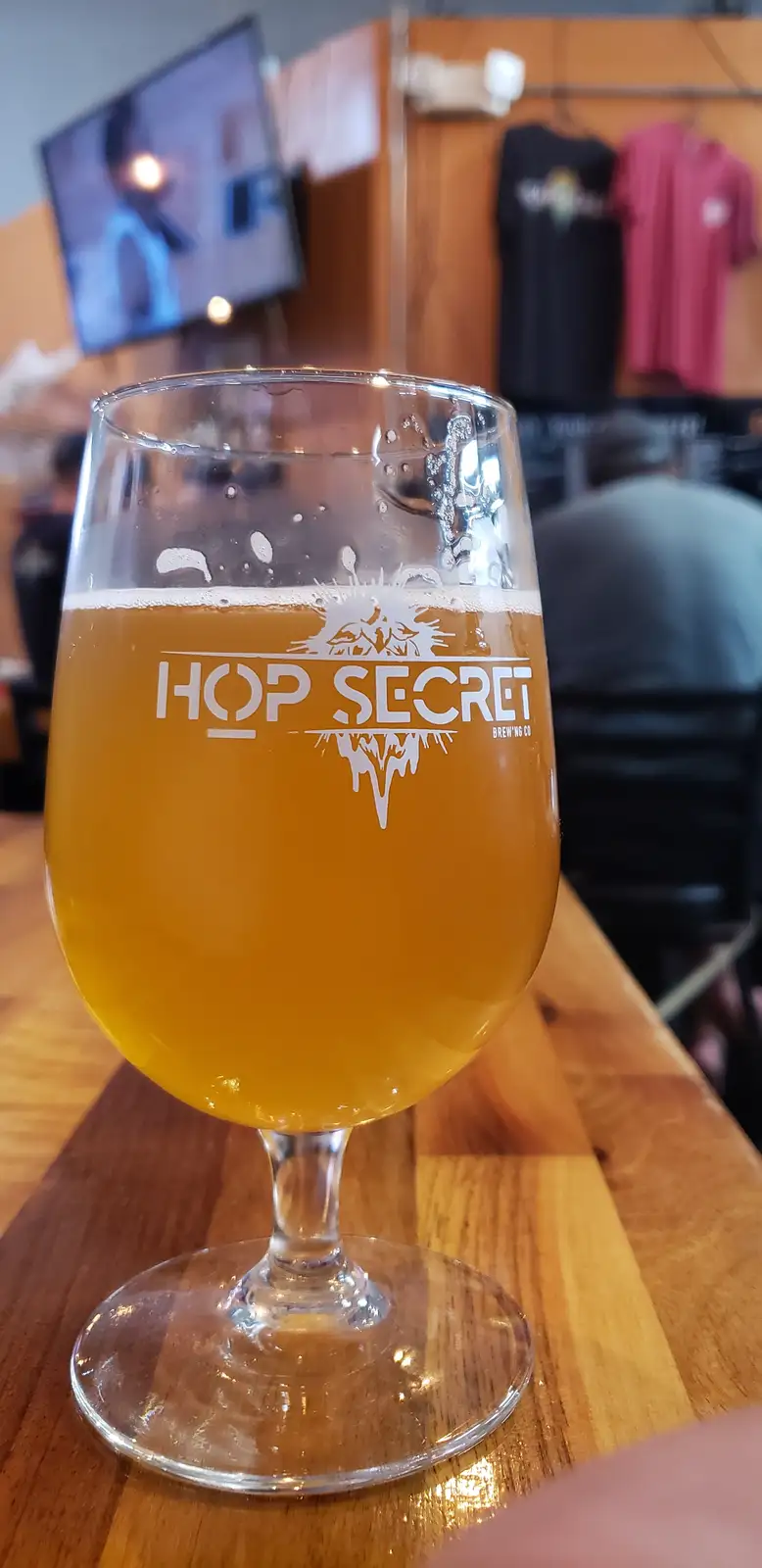 A close-up photo shows a stemmed beer glass filled with amber-colored beer and a white head, sitting on a wooden bar. The…