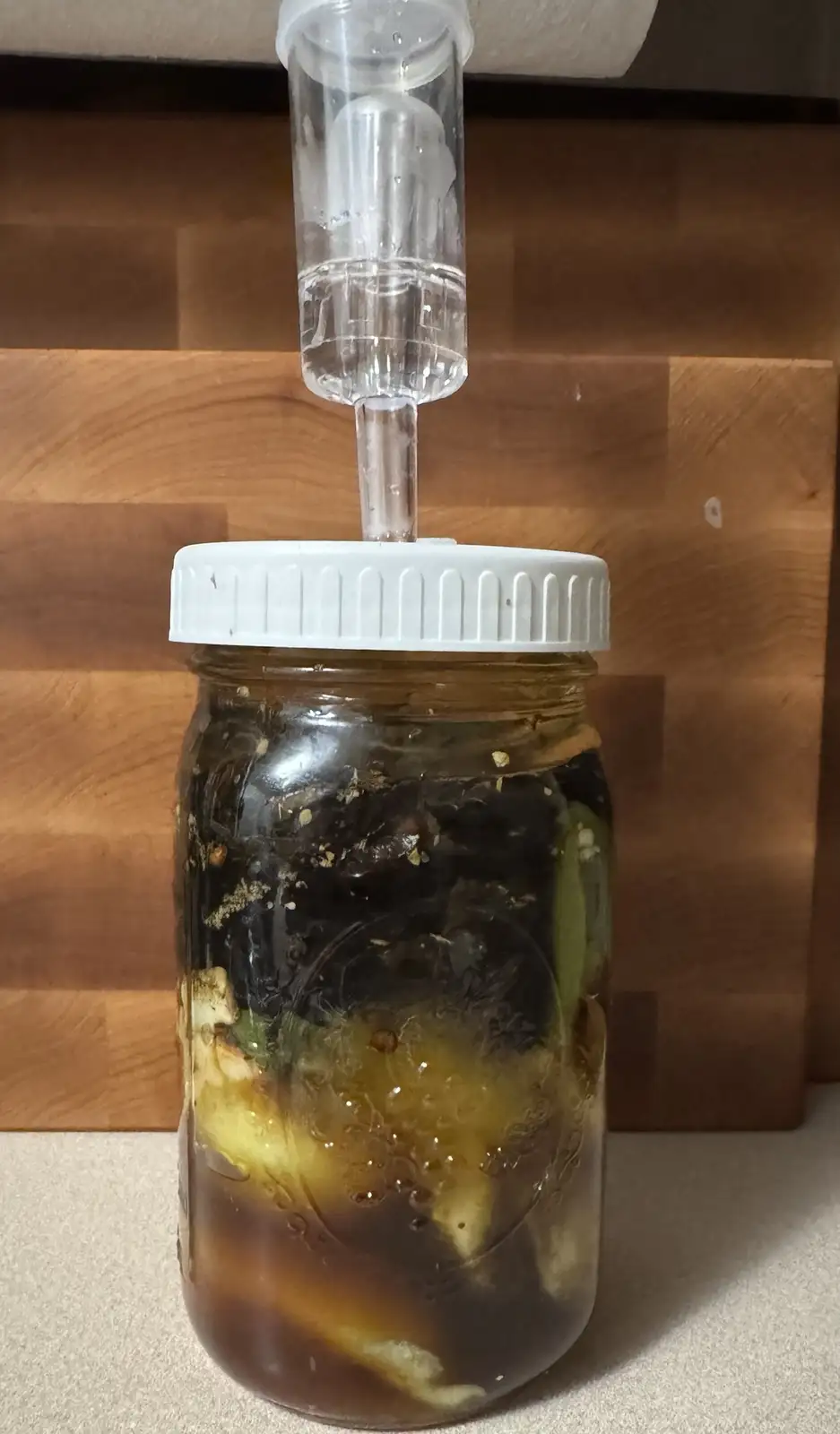 A glass mason jar filled with dark brine and pickled vegetables sits on a countertop, topped with a white plastic lid and a…
