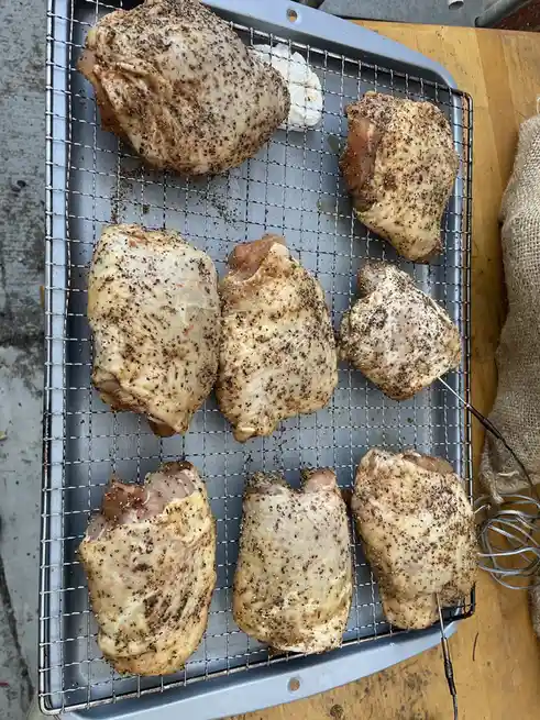 An overhead photo shows eight raw, seasoned chicken thighs arranged on a metal wire rack set over a baking sheet. The…