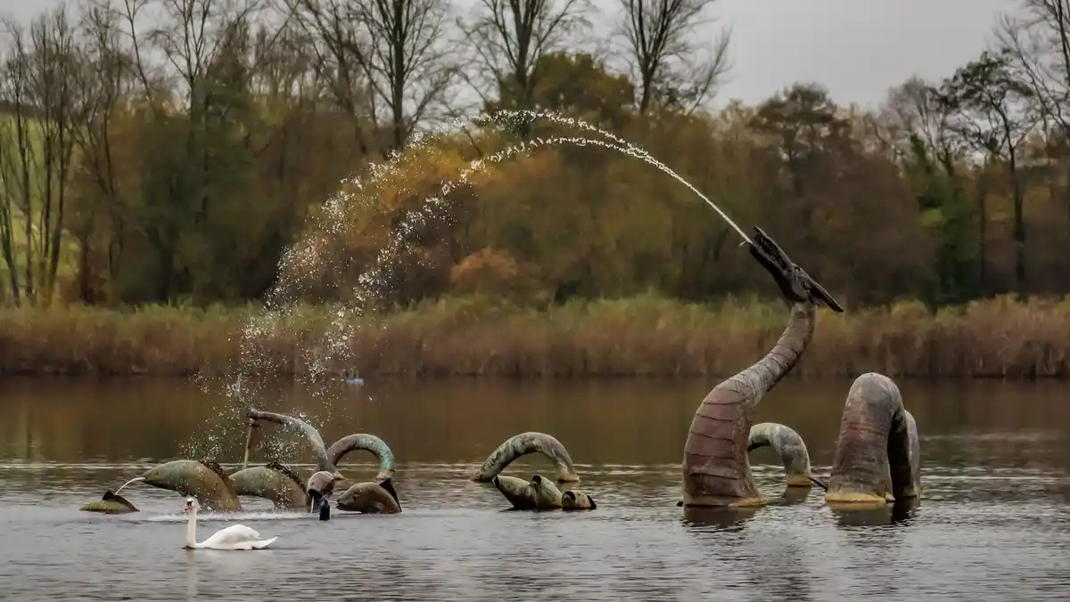 A serene lake scene features a white swan swimming calmly in the foreground, while multiple large, dark, dragon-like statues…