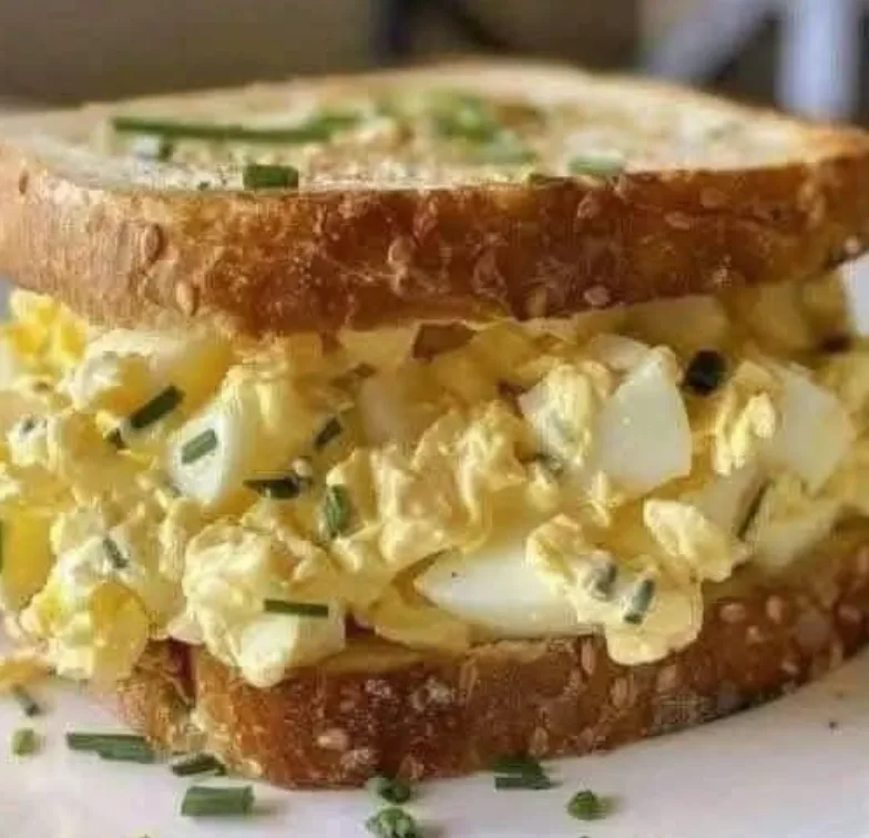 A close-up photo of a sandwich made with two slices of toasted whole grain bread, filled with creamy yellow egg salad mixed…