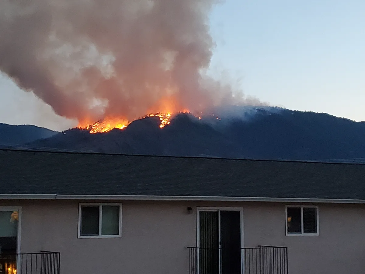 A photograph shows a large wildfire burning on a distant mountain ridge, with thick smoke rising into the sky, while the…
