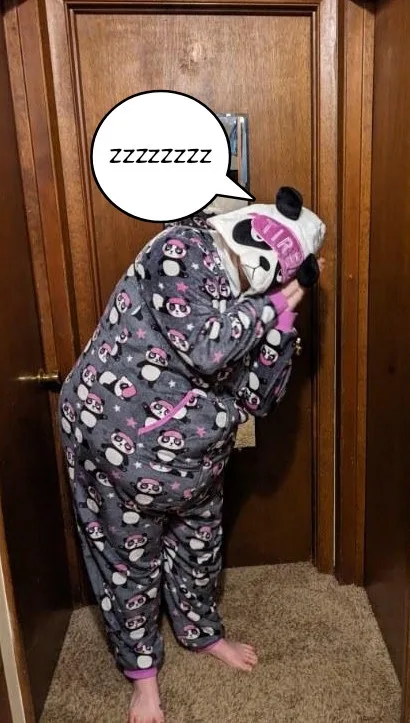 A person in a grey panda-themed onesie with pink accents is bent over, pressing their face against a wooden door, with a…