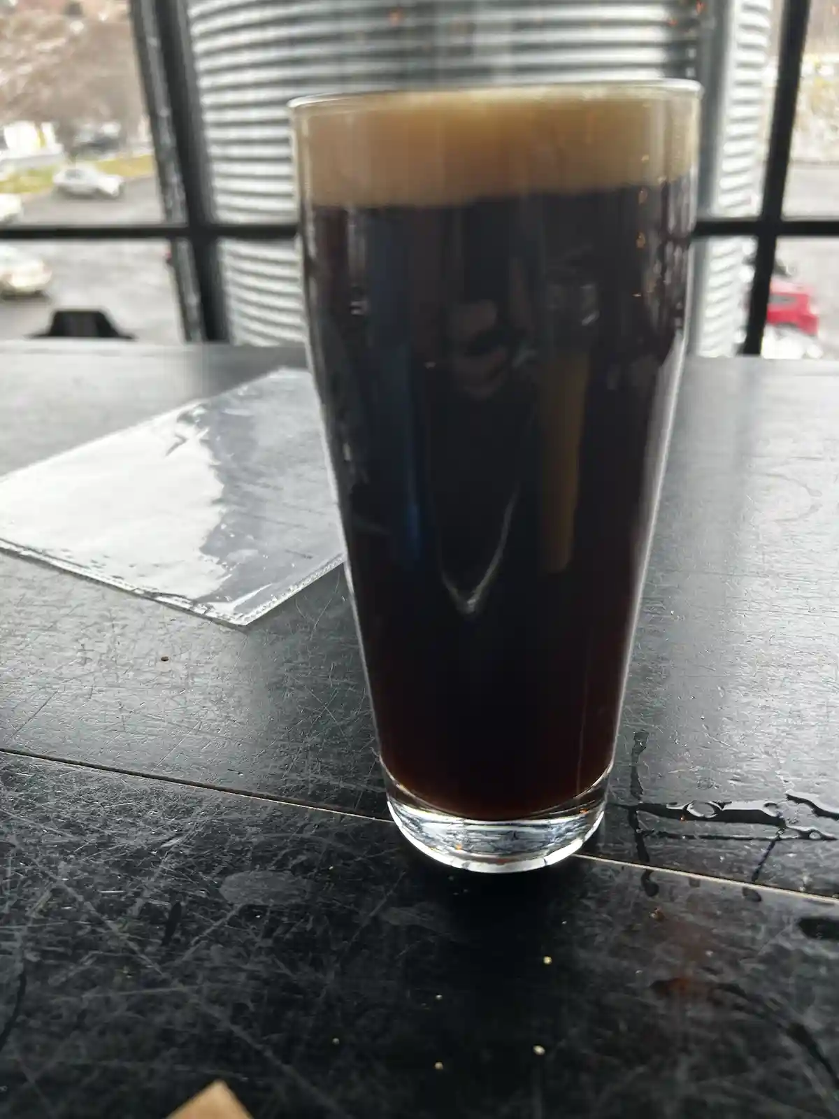 A tall glass of dark beer with a thick tan head sits on a scratched black table near a window. The overlaid text reads 'i'm…
